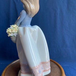 May include: A porcelain figurine of a girl in a blue dress and white skirt holding a basket of flowers. The figurine is standing on a wooden base.