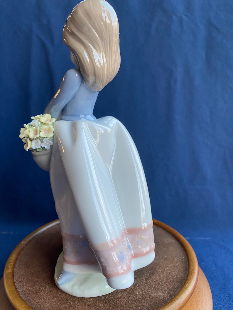 May include: A porcelain figurine of a girl in a blue dress and white skirt holding a basket of flowers. The figurine is standing on a wooden base.