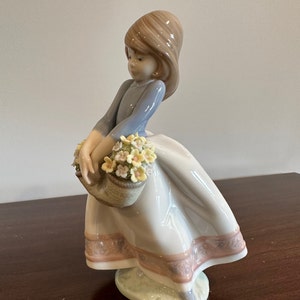 May include: A porcelain figurine of a girl in a blue dress and white skirt holding a basket of yellow flowers. The figurine is standing on a green base.