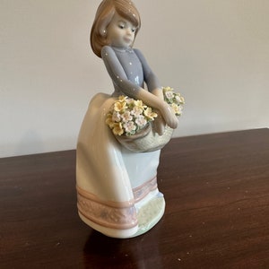 May include: A porcelain figurine of a girl in a blue dress and white skirt holding a basket of yellow and white flowers.