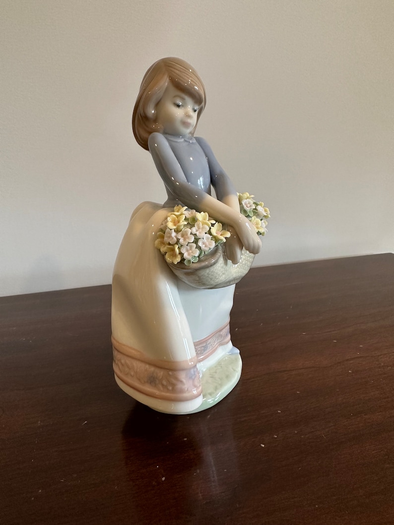 May include: A porcelain figurine of a girl in a blue dress and white skirt holding a basket of yellow and white flowers.