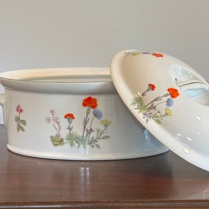 Può includere: Piatto da forno ovale in ceramica bianca con coperchio. Il piatto ha un design floreale con fiori arancioni, blu e verdi. Il coperchio ha un design floreale con fiori arancioni e verdi.