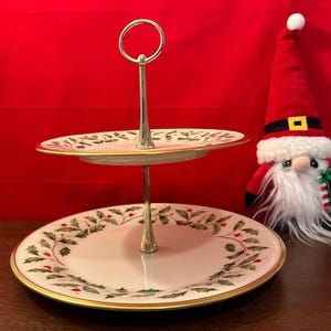 Può includere: Un portavivande a due livelli con un supporto argentato e piatti bianchi con un motivo di agrifoglio verde e bacche rosse. I piatti hanno un bordo dorato.