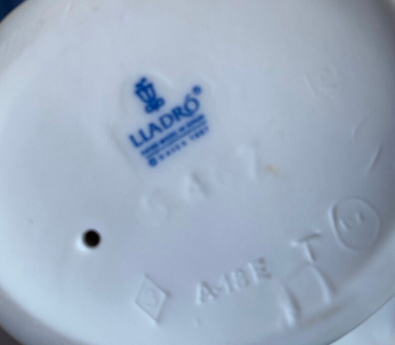 May include: A white porcelain figurine with a blue Lladro logo and the number 1477. The figurine is marked with the text "Made in Spain" and a diamond shape.