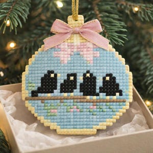 May include: A cross-stitch Christmas ornament in a round shape, featuring three black birds perched on a brown branch. The ornament has a light blue background with a pink bow and a gold hanging loop. The ornament is in a box.