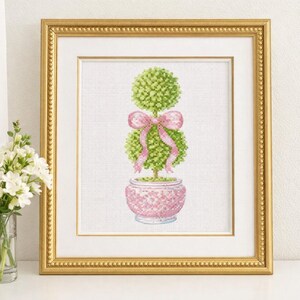 May include: Cross-stitch artwork of a topiary tree in a pink pot, adorned with a pink bow. The framed piece has a gold-colored frame. The artwork is displayed on a white surface, with a vase of white flowers to the left.