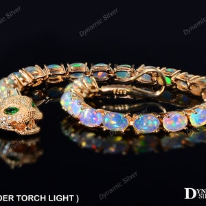 Puede incluir: Un brazalete de tono dorado con un cierre de cabeza de serpiente y piedras de ópalo ovaladas. El brazalete se muestra bajo una luz de antorcha, destacando los colores iridiscentes de los ópalos.