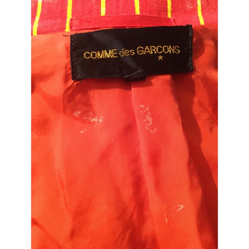Op de afbeelding: Close-up van de binnenkant van een rode jas met gele strepen. De voering is fel oranje van kleur. Een zwart label met gouden tekst "COMME des GARCONS" is in de jas genaaid.