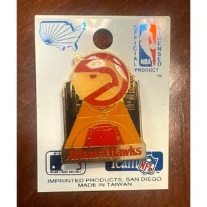 May include: An Atlanta Hawks team pin with the NBA official licensed product logo. The pin features the team's logo, a stylized hawk, and the words "Atlanta Hawks Team". The pin is on a white card with the MLB and NFL logos.