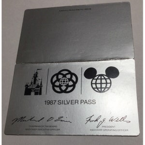 May include: A silver 1987 Silver Pass for Walt Disney World. The pass features the Walt Disney World logo, the Epcot logo, and the Mickey Mouse logo. The pass is signed by Michael D. Eisner, Chairman of the Board and Chief Executive Officer, and Frank G. Wells, President and Chief Operating Officer.