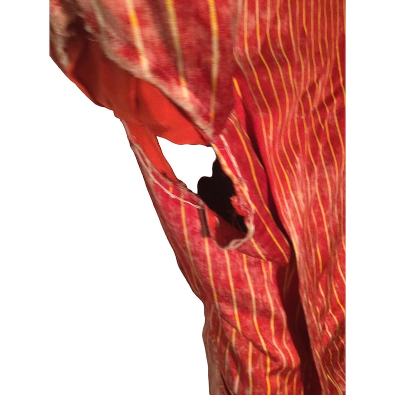 Op de afbeelding: Close-up van een rood kledingstuk met verticale gele strepen. De stof lijkt versleten en verweerd, met een zichtbare scheur die een oranje voering onthult. De textuur suggereert een zacht, mogelijk fluwelen materiaal.