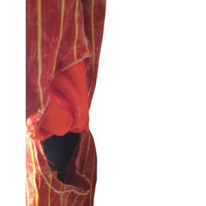 Op de afbeelding: Een rood kledingstuk met verticale gele strepen. De stof lijkt gedrapeerd te zijn en onthult een glimp van een effen oranje stof eronder. Het kledingstuk heeft een gescheurd gedeelte, waardoor een donker interieur zichtbaar wordt.