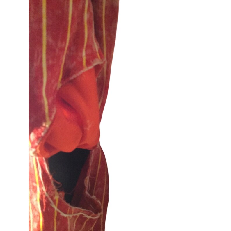 Op de afbeelding: Een rood kledingstuk met verticale gele strepen. De stof lijkt gedrapeerd te zijn en onthult een glimp van een effen oranje stof eronder. Het kledingstuk heeft een gescheurd gedeelte, waardoor een donker interieur zichtbaar wordt.