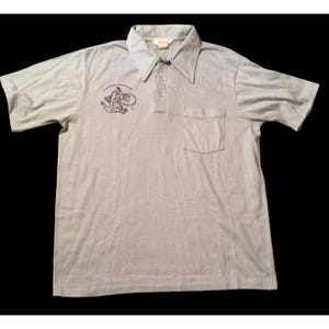 以下が含まれることがあります： 半袖、襟、胸ポケットが付いたライトグレーのポロシャツ。左胸には小さな刺繍のデザインとテキストがあります。シャツには3つのボタンと襟にタグが付いています。