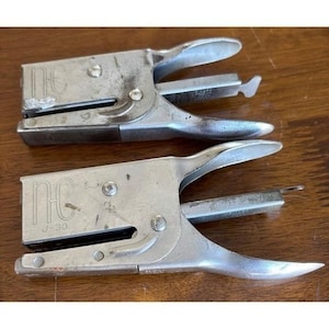 May include: Two vintage silver metal staplers. The staplers have a curved handle and a flat base. The text "NC" and "U-30" are visible on one of the staplers. The staplers are on a wooden surface.