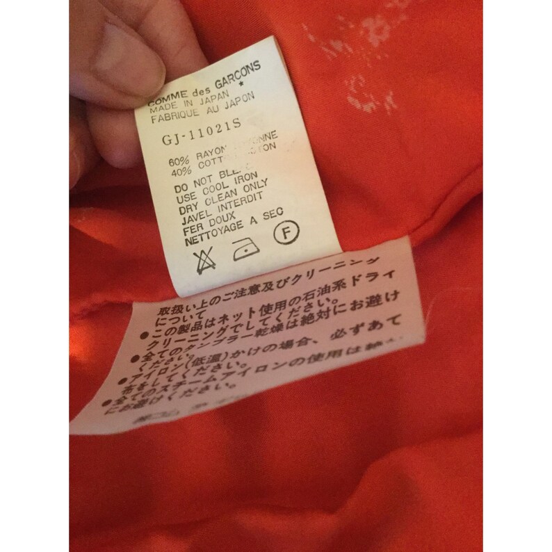 Op de afbeelding: Een close-up van een wit kledinglabel van Comme des Garcons, gemaakt in Japan. Zwarte tekst geeft de stof samenstelling (60% rayon, 40% katoen) en wasvoorschriften weer. De achtergrond is een feloranje stof.