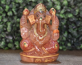 May include: A carved rose quartz Ganesha figurine featuring gold-painted accents on the crown, jewelry, and clothing. The deity is depicted in a seated position with an elephant head, large ears, and four arms, resting on a circular base.