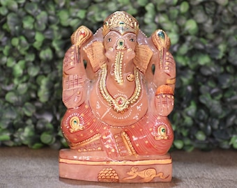 May include: A carved pink stone figurine of the deity Ganesha, featuring gold-painted accents on the crown, jewelry, and clothing. The figure is seated on a rectangular base with a small carved mouse and a bowl of sweets depicted on the front.