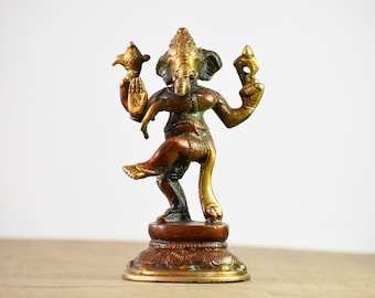 May include: A bronze-toned metal figurine of the deity Ganesha depicted in a dancing pose. The figure features four arms, an elephant head, and intricate engravings on the body, standing on a circular, ornate pedestal base with a dark finish.