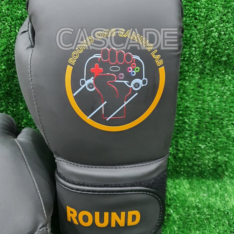 May include: A pair of black boxing gloves with a yellow and red logo that says "Round One Gaming Lab" and "Cascade". The gloves also have the word "Round" printed in yellow on the bottom.