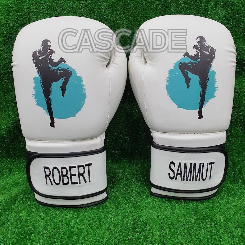 May include: A pair of white boxing gloves with black trim. The gloves feature a blue and white graphic of a person kicking. The gloves are labelled "Robert" and "Sammut".