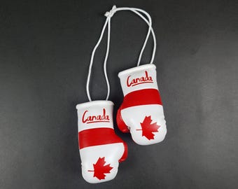 Op de afbeelding: Een paar miniatuur bokshandschoenen verbonden door een wit koord. De handschoenen zijn wit met een horizontale rode streep en een rood esdoornblad-embleem. Het woord Canada staat in rode letters op het bovenste witte gedeelte.