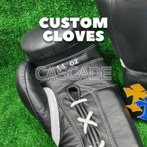 K&ouml;nnte beinhalten: Schwarze Leder-Boxhandschuhe mit wei&szlig;en N&auml;hten und Schn&uuml;rsenkeln. Die Handschuhe sind mit "14 oz" und "CASCADE" beschriftet. Der Text "CUSTOM GLOVES" wird &uuml;ber den Handschuhen angezeigt. Der Hintergrund ist gr&uuml;n.