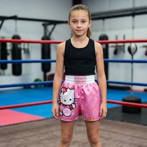 May include: A black sleeveless tank top paired with pink satin boxing shorts featuring a large Hello Kitty graphic, a white elastic waistband with the text Kitty Boxing, and a pattern of small white hearts and orange flowers on the fabric.