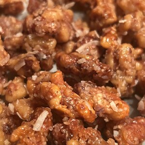 Könnte beinhalten: Eine Nahaufnahme von kandierten Walnüssen, die mit einer glänzenden, goldbraunen Zuckerglasur überzogen und mit kleinen, dünnen weißen Flocken aus Kokosraspeln bestreut sind. Die Textur der Nüsse ist uneben, zerklüftet und glänzend.