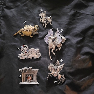 May include: Six silver-toned metal brooches featuring various designs. The brooches include a carousel horse, a mermaid with pearls, a Ferris wheel, a fireplace with a cat, and two winged horses.