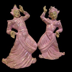 May include: Two ceramic figurines of dancers in pink and gold costumes. The figures are posed mid-dance, with arms raised and flowing skirts. The figurines have a vintage aesthetic, with gold accents and a glossy finish. The background is black.
