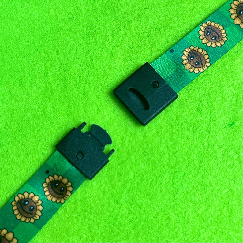 May include: Green lanyard with a repeating pattern of smiling sunflowers in yellow, brown, and green. The lanyard has a black plastic clasp and buckle. The background is a bright green fabric.
