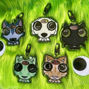 May include: Five acrylic keychains featuring stylized animal heads with oversized black eyes and small metal lobster clasps. The designs include a blue wolf, a white dog, a black creature, a light green animal, and an orange tabby cat, each with a small cutout.