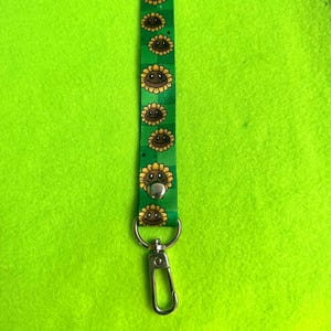 May include: Green lanyard with a repeating pattern of smiling sunflowers. The lanyard has a silver-colored metal clasp and a snap closure. The background is a bright green.