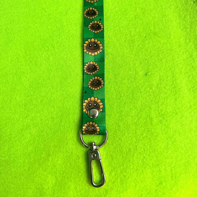 May include: Green lanyard with a repeating pattern of smiling sunflowers. The lanyard has a silver-colored metal clasp and a snap closure. The background is a bright green.