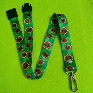 May include: Green lanyard with a repeating pattern of smiling sunflowers. The lanyard has a metal clasp and a black plastic clip. The background is a solid, bright green.