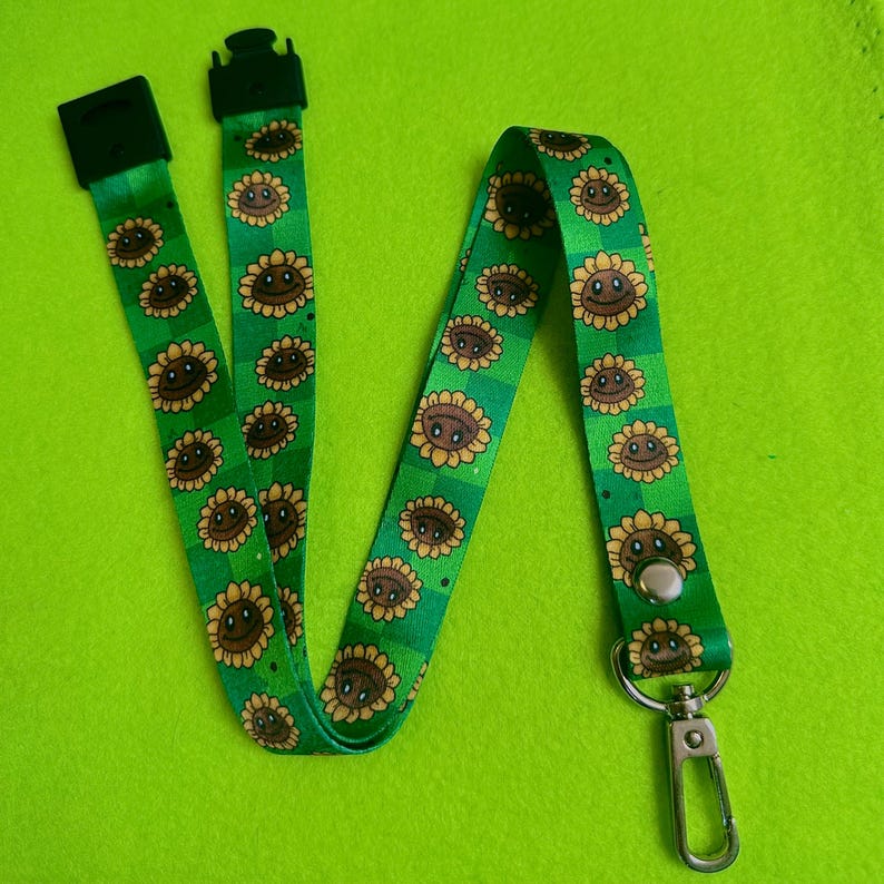 May include: Green lanyard with a repeating pattern of smiling sunflowers. The lanyard has a metal clasp and a black plastic clip. The background is a solid, bright green.