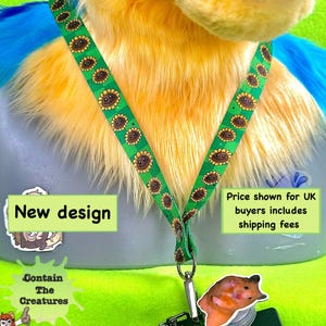 Op de afbeelding: Groene lanyard met een zonnebloemmotief en een hamsterbedel. De lanyard is om een ​​pluche speelgoed met gele, blauwe en witte vacht. De tekst "New design" is zichtbaar op een sticker.