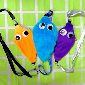 Peut inclure: Trois sacs à bandoulière originaux en forme de ver, orange, bleu et violet. Chaque sac est orné de grands yeux globuleux et d'une sangle assortie. Les sacs sont suspendus sur un fond à carreaux vert et blanc.