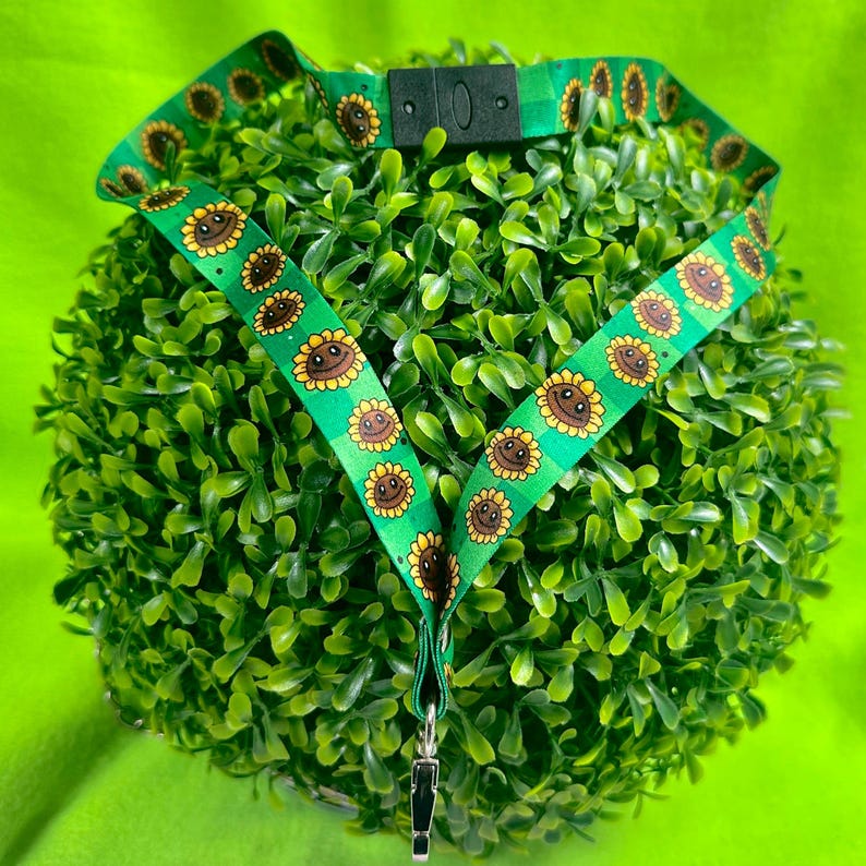 May include: Green lanyard with a repeating pattern of smiling sunflowers. The lanyard has a black plastic clasp and a metal clip at the bottom. The sunflowers are yellow and brown with green stems and leaves.