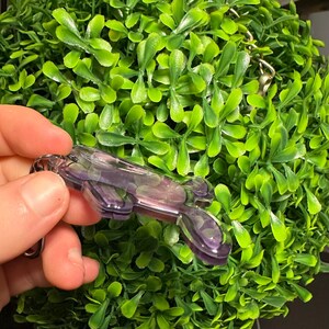 May include: A purple and clear resin keychain with a silver-colored chain. The keychain is held in front of a green, leafy, spherical plant. The resin has a marbled appearance with shades of purple and clear.