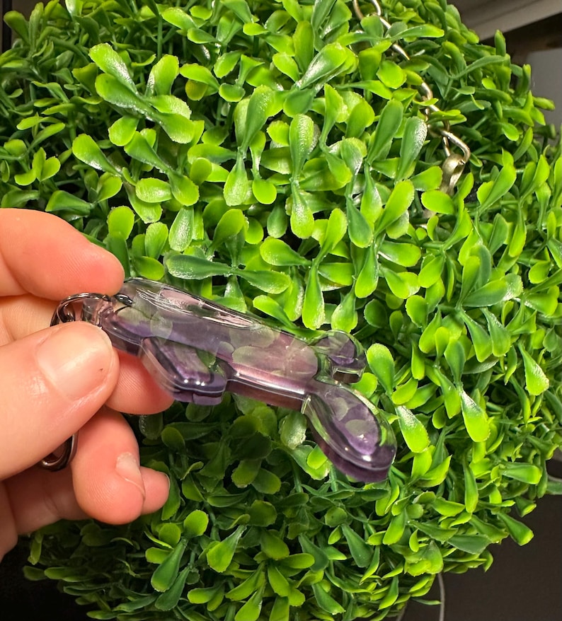 May include: A purple and clear resin keychain with a silver-colored chain. The keychain is held in front of a green, leafy, spherical plant. The resin has a marbled appearance with shades of purple and clear.