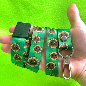 May include: Green lanyard with a repeating pattern of smiling sunflowers. The lanyard has a black plastic clasp and a silver-colored metal clip for attaching keys or an ID badge. The background is a solid, bright green.