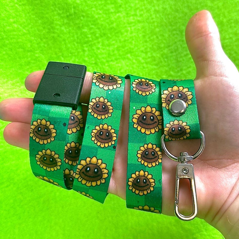 May include: Green lanyard with a repeating pattern of smiling sunflowers. The lanyard has a black plastic clasp and a silver-colored metal clip for attaching keys or an ID badge. The background is a solid, bright green.
