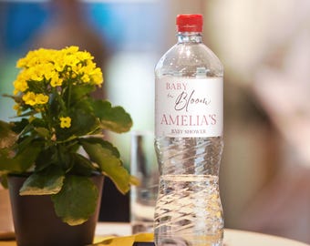 Könnte beinhalten: Eine transparente Plastikwasserflasche mit rotem Deckel und einem Etikett mit der Aufschrift "Baby in Bloom, Amelia's, Baby Shower". Im Hintergrund befindet sich eine Topfpflanze mit gelben Blüten und grünen Blättern.