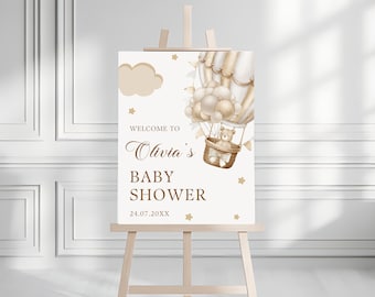 May include: A white baby shower welcome sign with a brown teddy bear in a hot air balloon. The balloon is filled with gold and white balloons and has gold stars and a cloud in the background. The sign says "Welcome to Olivia's Baby Shower 24.07.20XX".