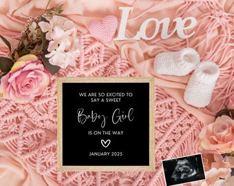May include: A black chalkboard sign with white text announcing the arrival of a baby girl in January 2025. The sign is surrounded by pink crocheted fabric, white booties, and pink flowers.