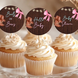 Puede incluir: Cupcakes con glaseado blanco y adornos decorativos. Los adornos son redondos con un fondo de vetas de madera, botas de vaquero, lazos rosas y el texto "Boots or Bows", "Hello Baby" y "Baby Shower".