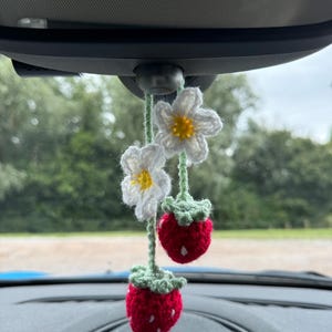 May include: A crocheted strawberry and flower car charm hanging from the rear view mirror of a car. The charm is made of red, white and green yarn.