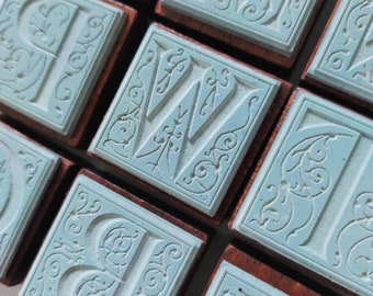 May include: A set of wooden alphabet stamps with intricate, decorative designs. Each stamp features a different letter in a light blue color, surrounded by a detailed floral pattern. The stamps are perfect for adding a vintage touch to crafts and projects.